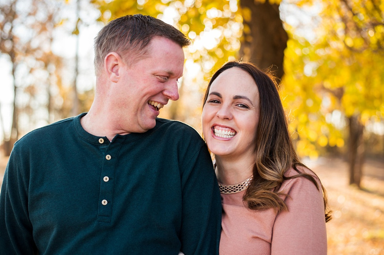 a husband and wife are laughing together, while he looks at her and she looks at the camera with bright fall color behind them at a park in moore, oklahoma at a family photo session