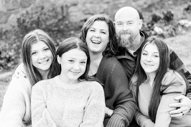 Black and white image of A mom and dad with 3 teen daughters sit and smile together on the rocks with fall color around them at Will Rogers Gardens in Oklahoma City, Oklahoma