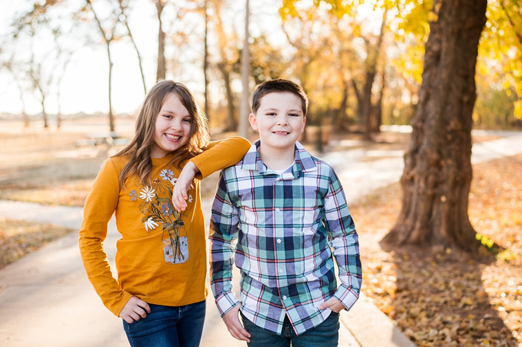 a brother and sister stand together, with her elbow on his shoulder and smile at a family photo session in the fall at a park in Moore, Oklahoma