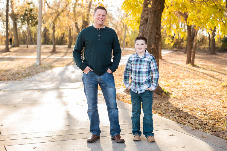 a father and sound stand the same with hands in pockets and smile on a sidewalk at a park in moore, Oklahoma with fall color behind them at a family photo session