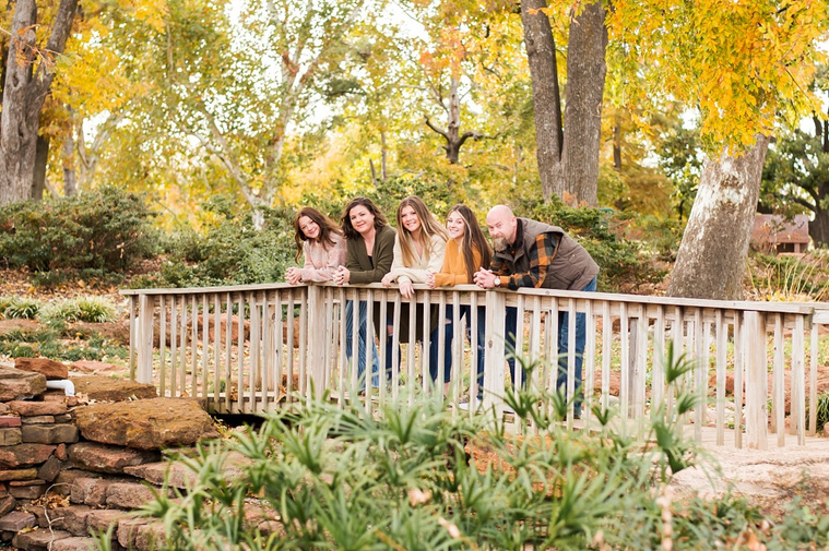 A mom and dad with 3 teen daughters stand on a wooden footbridge and smile together with fall color around them at Will Rogers Gardens in Oklahoma City, Oklahoma
