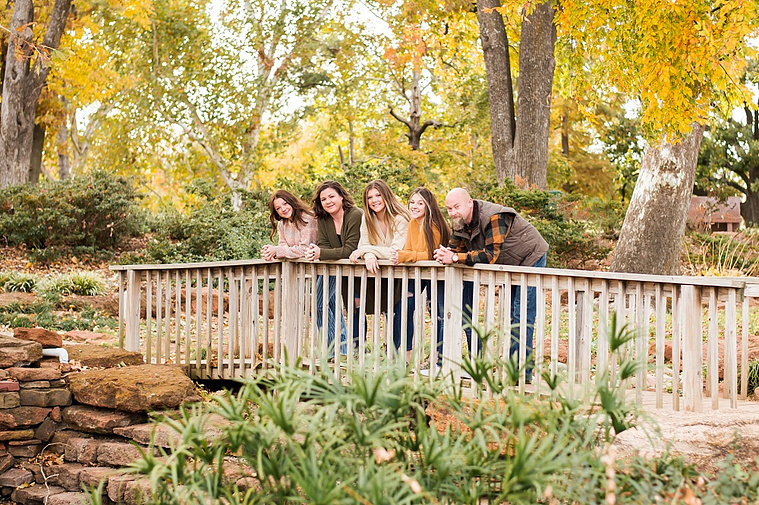 A mom and dad with 3 teen daughters stand on a wooden footbridge and smile together with fall color around them at Will Rogers Gardens in Oklahoma City, Oklahoma