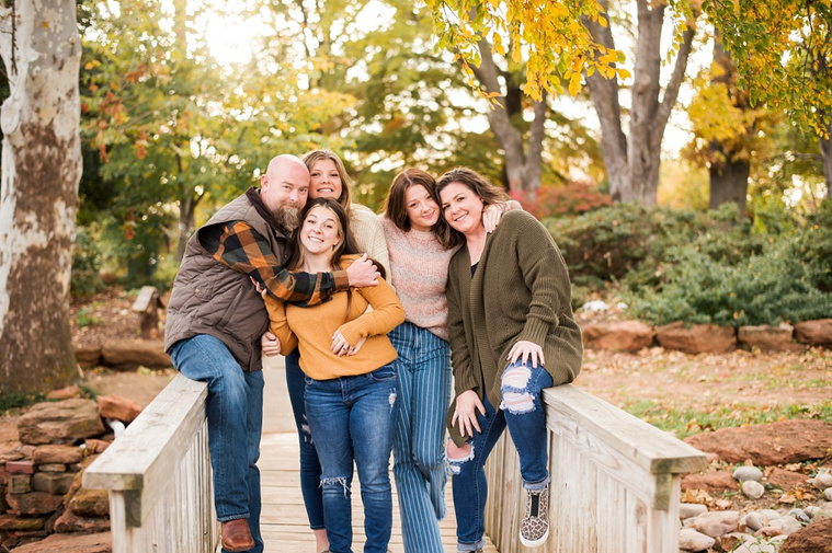 A mom and dad with 3 teen daughters stand on a wooden footbridge and smile together with fall color around them at Will Rogers Gardens in Oklahoma City, Oklahoma