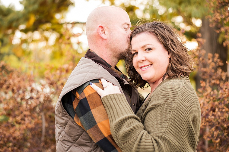 A husband and wife hug with the husband kissing her temple with fall color all around them at Will Rogers Gardens in Oklahoma City, Oklahoma