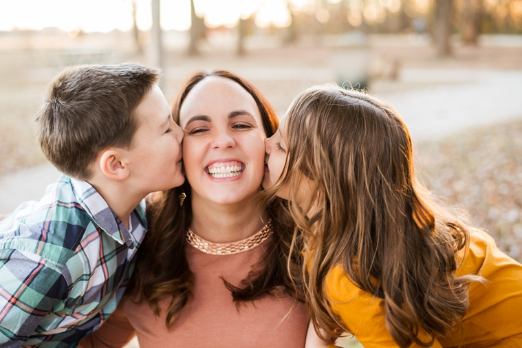 a son and daughter stand on either side of their mom and kiss her cheeks while she smiles at a family photo session in a park in Moore, Oklahoma in the fall