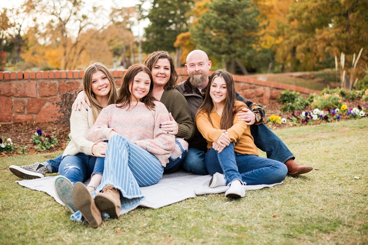 A mom and dad with 3 teen daughters sit and smile together on the grass with flowers and rock wall behind them, and with fall color around them at Will Rogers Gardens in Oklahoma City, Oklahoma