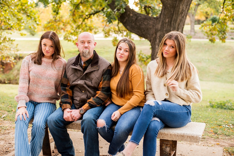 A dad with 3 teen daughters sit together on a bench and make goofy faces with fall color around them at Will Rogers Gardens in Oklahoma City, Oklahoma