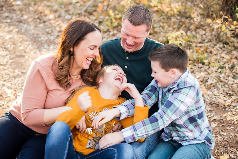 a family of four with a young boy and girl sit together on the ground in the woods and tickle the girl laughing together at a family photo session in Moore, Oklahoma