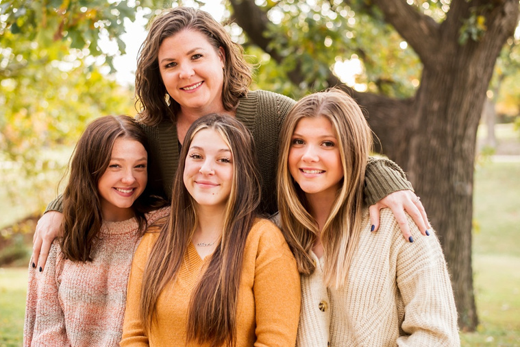A mom and 3 teen daughters sit and hug and smile together on a bench with fall color around them at Will Rogers Gardens in Oklahoma City, Oklahoma
