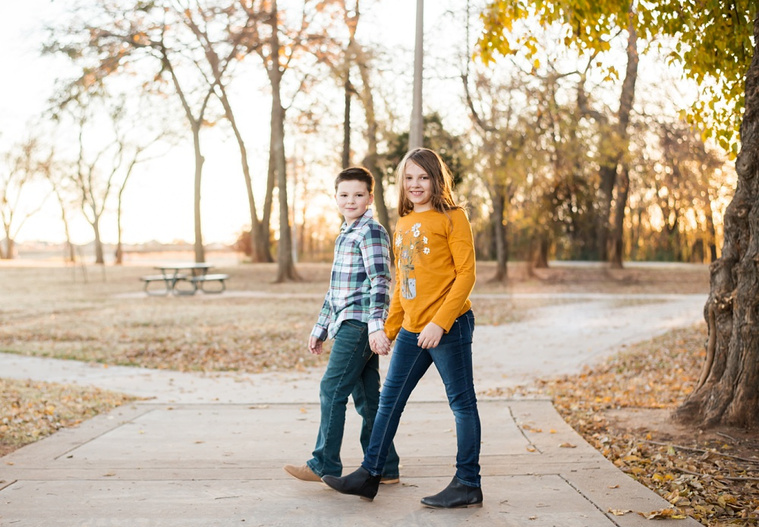 a brother and sister walk together across the frame holding hands and smiling on a sidewalk in a park in Moore, Oklahoma at a family photo session
