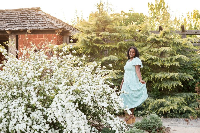 senior girl wearing a dress stands prettily and smiles holding her skirt out among the flowers and trees of Will Rogers Gardens in OKC