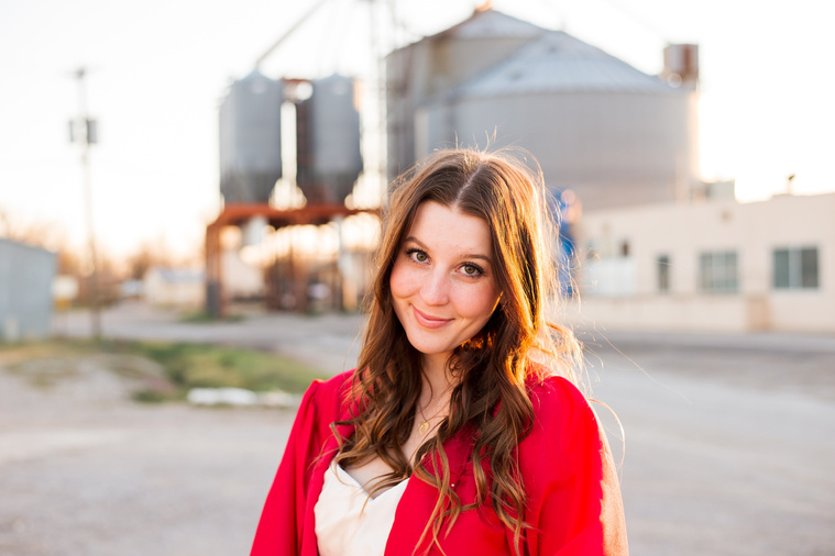 high school senior girl wearing a white dress and her red graduation gown smiles sweetly in front of the grainery in Tuttle Oklahoma