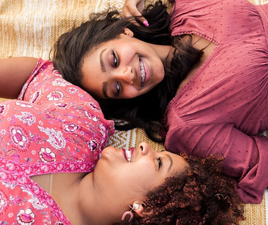 a senior girl and her mom lay on their backs with faces looking at each other and smiling on a tan rug in a field in central Oklahoma