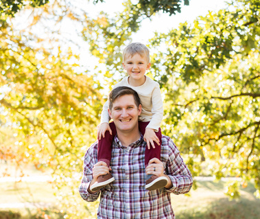 a father stands with this young son on his shoulders and smiles at Will Rogers Park in Oklahoma City, Oklahoma at a fall photo mini session by Cheryl Jackson Photography
