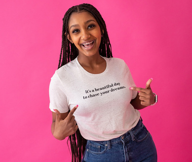 black girl with long weave stands in front of a pink studio backdrop wearing a graphic tee and blue jeans smiling and pointing, taken by Oklahoma City branding photographer