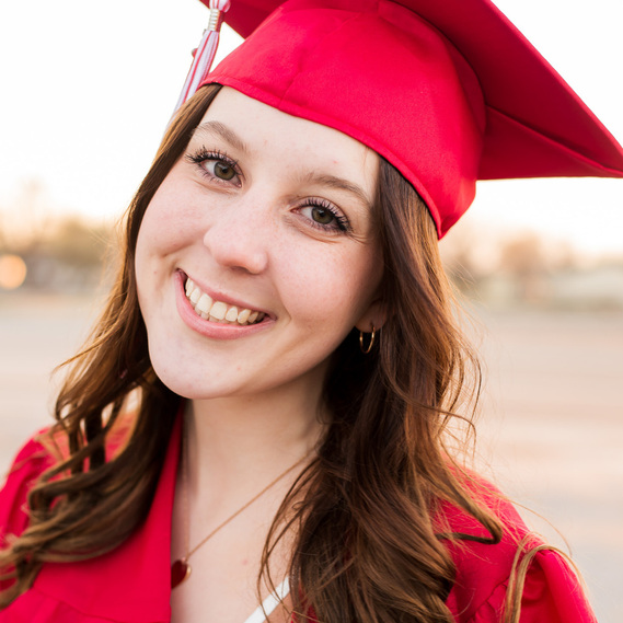high school senior girl with long brown hair smiles prettily wearing her red graduation cap, tassel and gown on a street in Tuttle Oklahoma