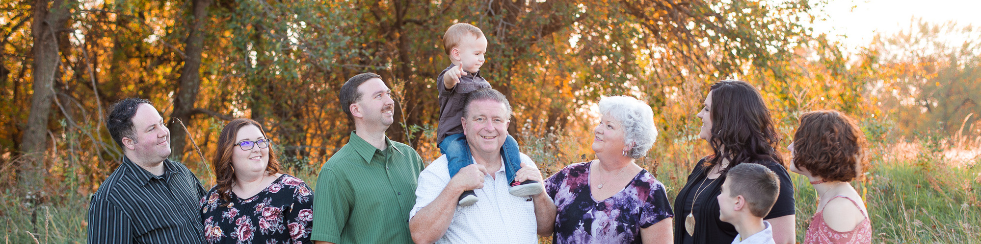 A multi-generational family portrait of a toddler on his grandpa's shoulders pointing at the camera and everyone else looking at him.