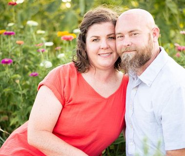 A couple sit together in front of a flower field near Oklahoma City, Oklahoma and smile at the camera with their cheeks touching.