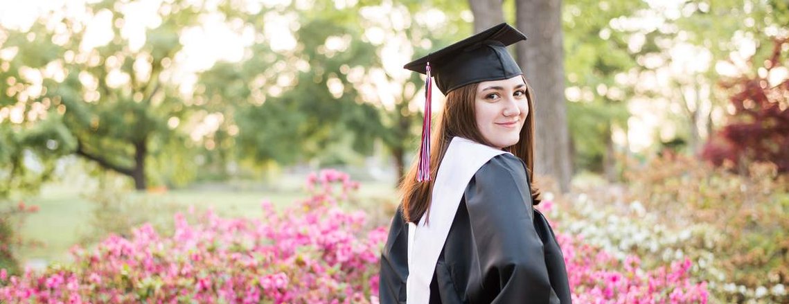 a high school senior girl wearing her graduation cap and gown poses with hands on hips and looking back over her shoulder in front of pink flowers at will rogers gardens in oklahoma city