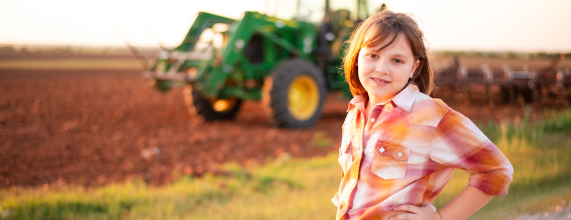A little girl smiles with her hand on her hip with a John Deere tractor in a plowed field behind her, at her back to school photo session in the country in Oklahoma