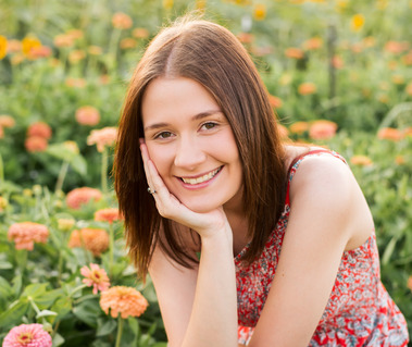 High school senior girl sits in the middle of a flower field in Oklahoma with her chin in her hands smiling 