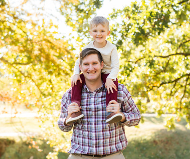 a dad with his toddler son on his shoulders smiling in front of trees at Will Rogers Gardens