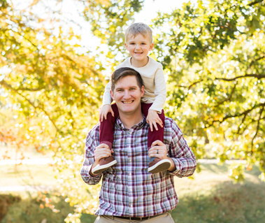a dad with his toddler son on his shoulders smiling in front of trees at Will Rogers Gardens
