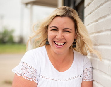 Cheryl Jackson, photographer laughs and smiles in a headshot leaning against a white brick wall in Oklahoma City, OK.