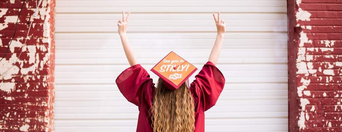 high school senior faces away from camera with cap and gown on holding up her fingers as twos on either side, cap has college sticker on it