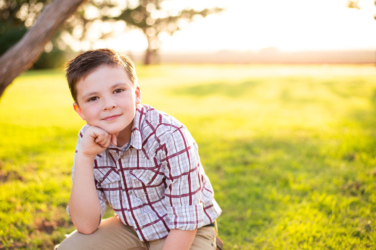 A little boy sits with his hand on his chin in a yard in his back to school photo session in the country in Oklahoma