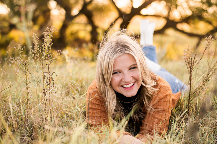 high school senior girl with blond hair wearing a rust colored sweater and jeans, lays on her stomach and laughs in a golden field in Oklahoma for her photo session