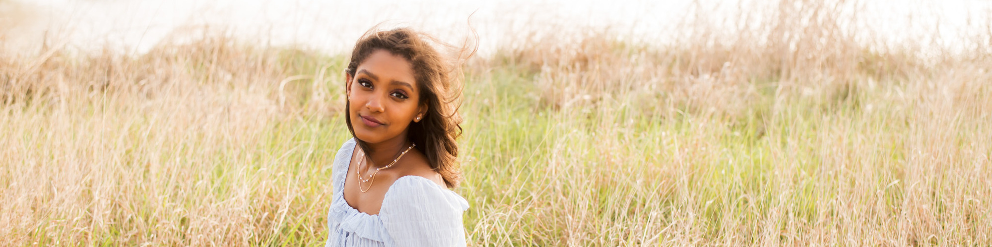 high school senior girl with dark skin and hair wearing a light blue dress sits in tall grass with a small pond behind her in a field in central Oklahoma
