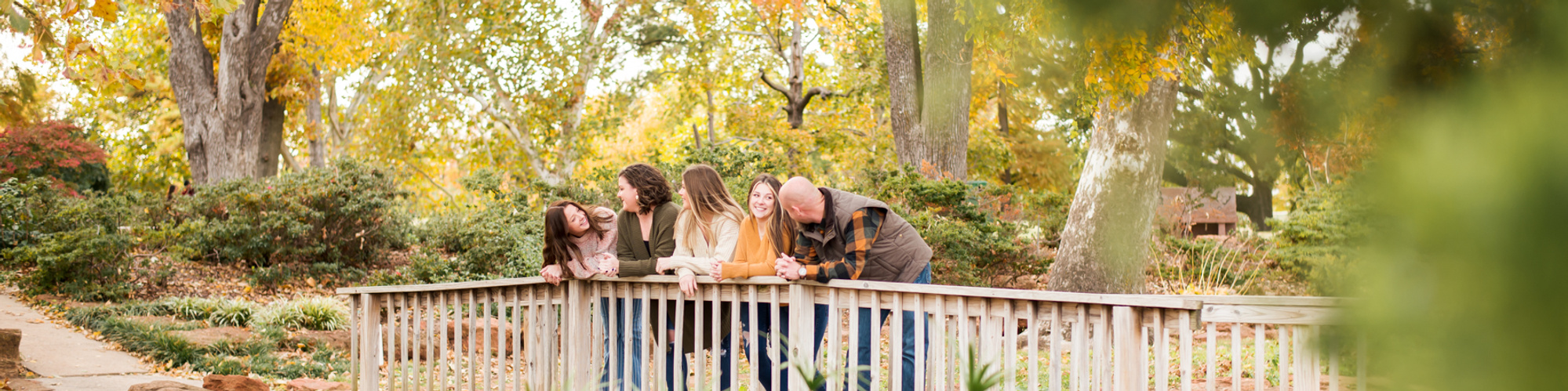 wide angle shot of a mom and dad with their 3 teenage daughters standing on a foot bridge and talking with fall foliage and trees all around them at Will Rogers Gardens in Oklahoma City