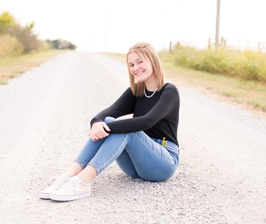 Portrait of a teenage girl in a black t-shirt and jeans sitting in the middle of a dirt road with her hands around her knees smiling at the camera
