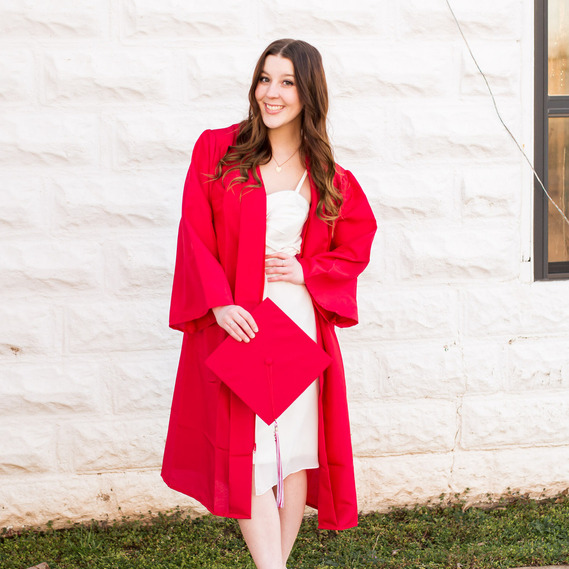 full portrait of a high school senior girl with long brown hair wearing a red graduation gown smiling and holding  her red cap with hand on her waist and white dress in front of a white brick wall in downtown Tuttle Oklahoma