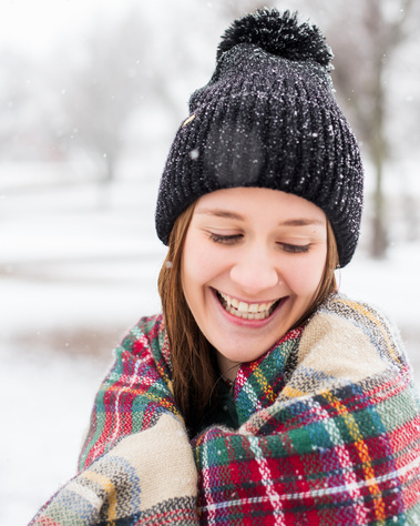 portrait of a high school senior girl wrapped in a red plaid blanket wearing a black beanie laughing while she stands in the snow in a park in Tuttle Oklahoma