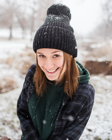 portrait of a high school senior girl wearing a black beanie and dark plaid jacket smiling up while she stands in the snow in a park in Tuttle Oklahoma