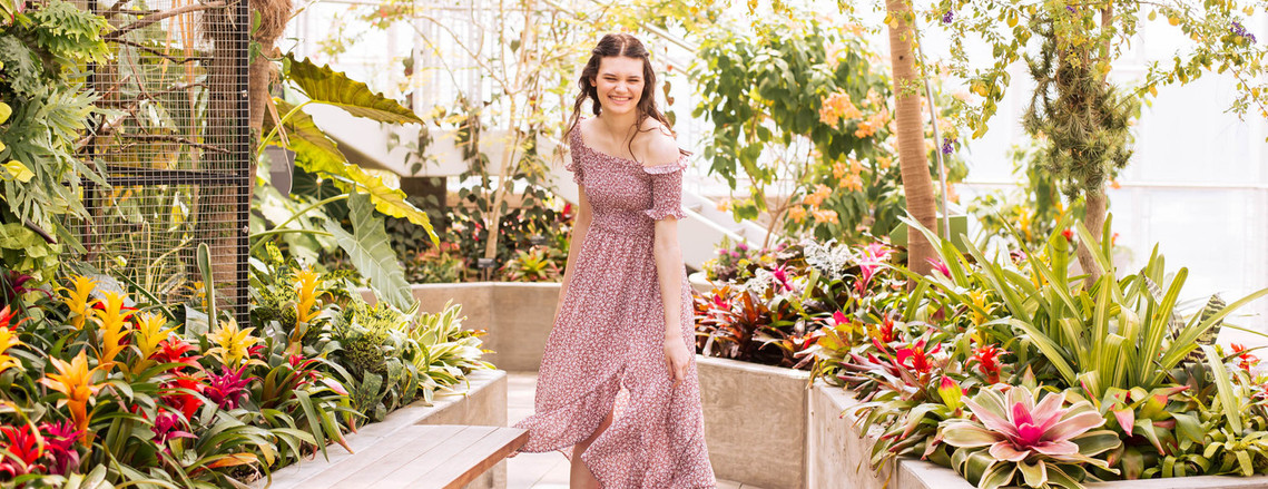 senior girl with long brown hair wearing a flowy red dress spins and laughs on a path with tropical flowers and plants around her in the Crystal Bridge at Myriad Botanical gardens in Oklahoma city, taken by OKC senior photographer