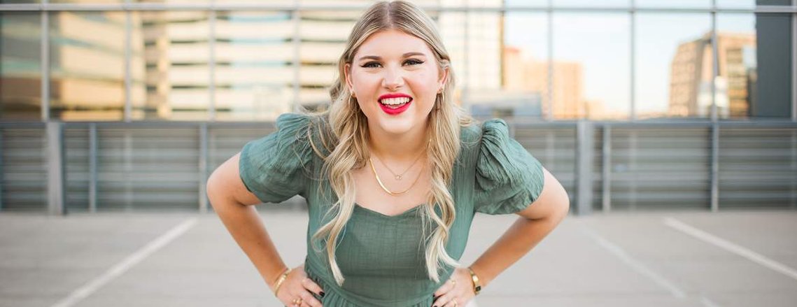 blond high school senior girl stands with her hands on her hips and smiles in a green dress in front of Devon tower on the parking garage rooftop in downtown Oklahoma City