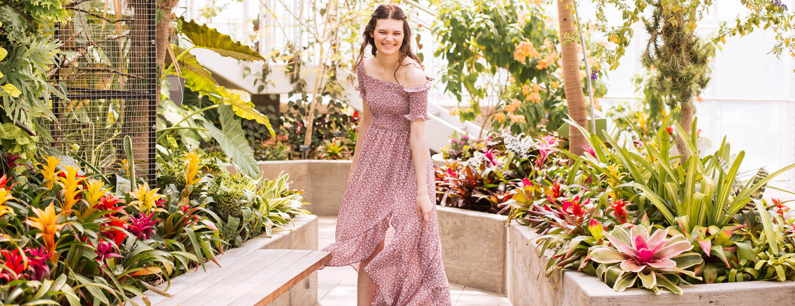 senior portrait of a girl wearing a patterned red flowy dress twirling and laughing among tropical plants and flowers in a walkway in the Crystal Bridge at Myriad Gardens in Oklahoma City, Oklahoma