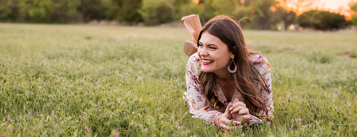 high school senior girl with long brown hair blowing lays on the grass on here stomach and propped on elbows while she laughs to the side at sunset in a field in south Oklahoma City taken by OKC senior photographer