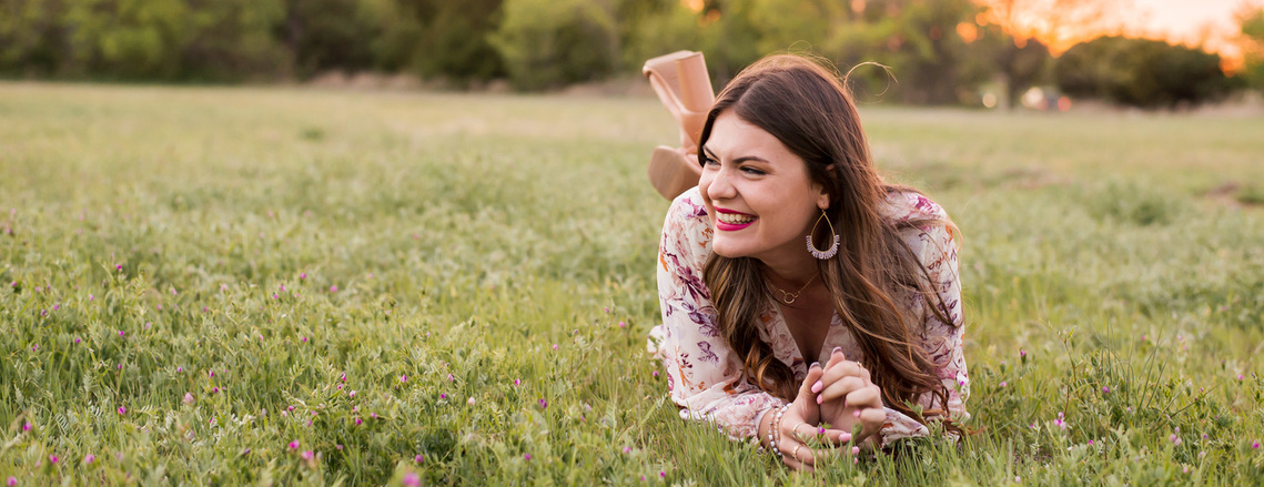 high school senior girl with long brown hair blowing lays on the grass on here stomach and propped on elbows while she laughs to the side at sunset in a field in south Oklahoma City