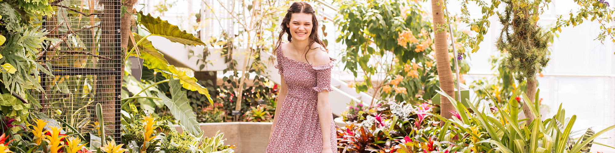 panoramic image of a senior girl with brown hair laughing and twirling in the middle of tropical plans and flowers inside Crystal Bridge at Myriad Gardens in downtown Oklahoma City