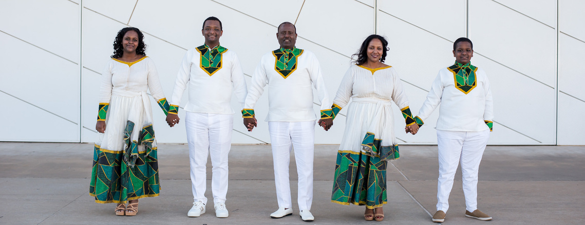 Mom and dad with 3 teenage children dressed in authentic Ethiopian attire stand apart and hold hands in front of the Devon Boathouse in Oklahoma City