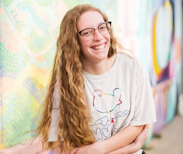 high school senior girl with long wavy blond hair wearing glasses hugs each elbow and smiles in front of a pastel colored mural at Factory Obscura in Oklahoma City