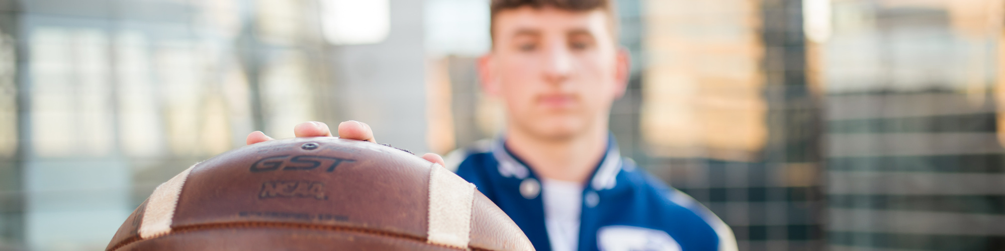 senior boy wearing a letterman jacket is slightly out of focus while he holds a football close to camera that is in focus with OKC downtown buildings behind him