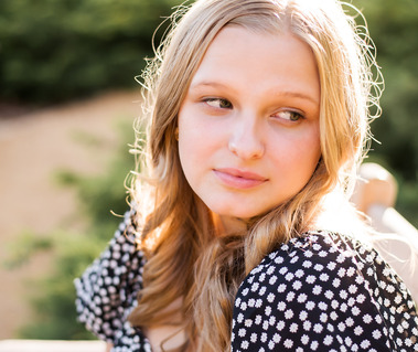 blond senior girl wearing a black and white dress sits on a bench and looks off to side softly with rim light on her head at Myriad Gardens, taken by Oklahoma City senior photographer