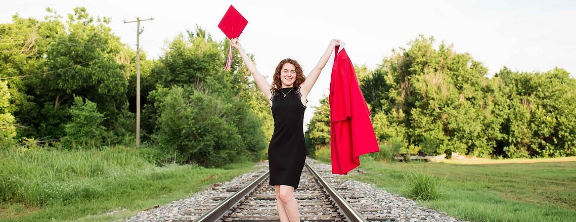 a senior girl wearing a black dress holds up her red graduation cap and gown in each hand with a big smile standing in the  middle of railroad tracks in Yukon, Oklahoma