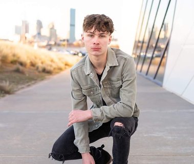 teen boy kneels on sidewalk next to Devon Boathouse in Oklahoma City with skyline behind him