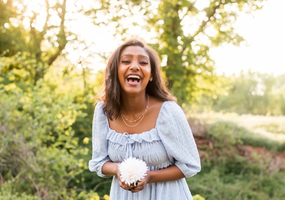 black senior girl wearing light blue dress holding a white flower laughs joyfully in front of trees and fields in Oklahoma